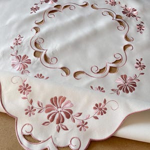May include: White tablecloth with a pink floral embroidery design. The embroidery features a scalloped border and a circular design in the center.