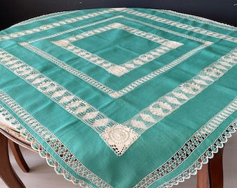 Könnte beinhalten: Eine tealfarbene Tischdecke mit einer weißen Spitzenbordüre und aufwendigen Stickereien. Die Tischdecke ist quadratisch und hat ein zartes, feminines Design.