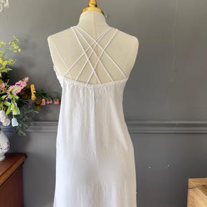 May include: A white sleeveless dress featuring a crisscross strap design on the back. The garment is displayed on a mannequin, showcasing a simple, solid white fabric construction with thin, delicate straps that form an intricate pattern across the upper back area.