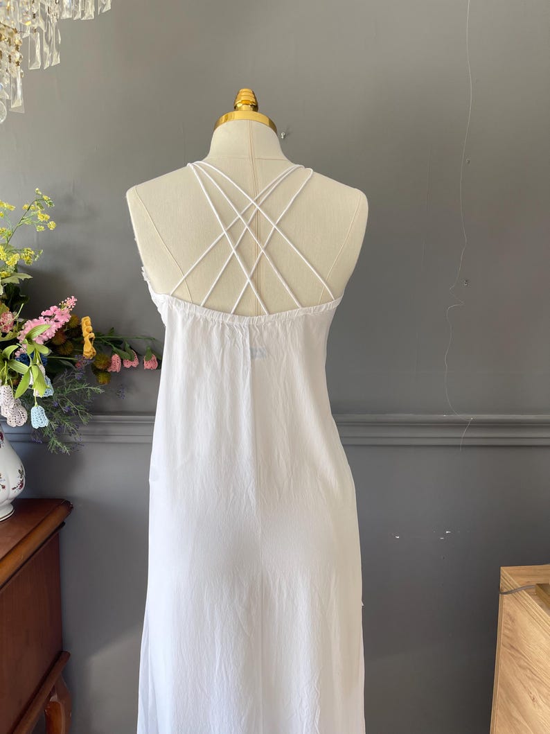 May include: A white sleeveless dress featuring a crisscross strap design on the back. The garment is displayed on a mannequin, showcasing a simple, solid white fabric construction with thin, delicate straps that form an intricate pattern across the upper back area.