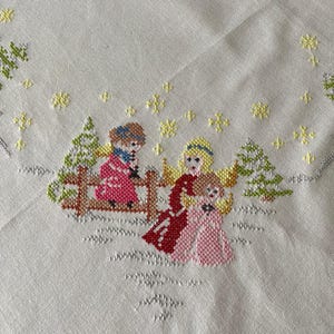 Puede incluir: Un mantel de lino blanco con un diseño de punto de cruz que representa a tres niños con ropa de invierno de pie frente a una valla con árboles y copos de nieve. Los niños visten ropa roja, rosa y azul.