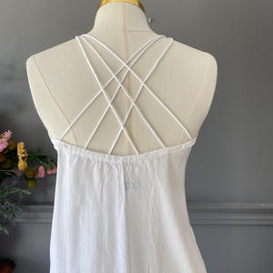 May include: A white sleeveless dress featuring a back design with multiple thin, crisscrossing straps that form a geometric pattern. The garment is displayed on a mannequin, showcasing the plain white fabric and the intricate strap arrangement.