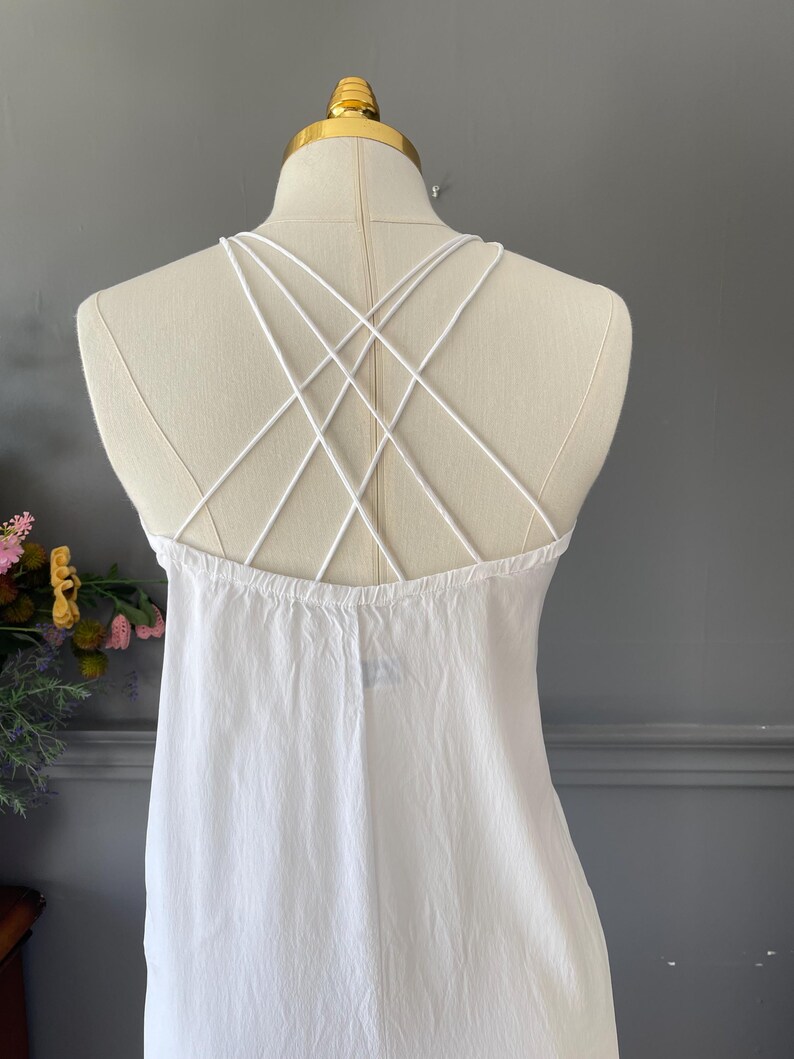 May include: A white sleeveless dress featuring a back design with multiple thin, crisscrossing straps that form a geometric pattern. The garment is displayed on a mannequin, showcasing the plain white fabric and the intricate strap arrangement.