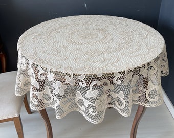 Könnte beinhalten: Eine weiße Häkeltischdecke mit einem zarten Blumenmuster. Die Tischdecke ist rund und hat einen gewellten Rand. Sie ist über einen Holztisch gelegt.
