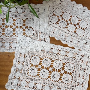 Puede incluir: Tres tapetes rectangulares blancos de ganchillo con un patrón floral y bordes festoneados se exhiben sobre una superficie de madera. Los tapetes muestran un intrincado diseño calado, perfecto para la decoración del hogar.