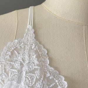 May include: A white camisole featuring delicate floral lace trim along the neckline and thin, triple-strand spaghetti straps. The garment is displayed on a beige mannequin, highlighting the intricate embroidery and scalloped edges of the lace.