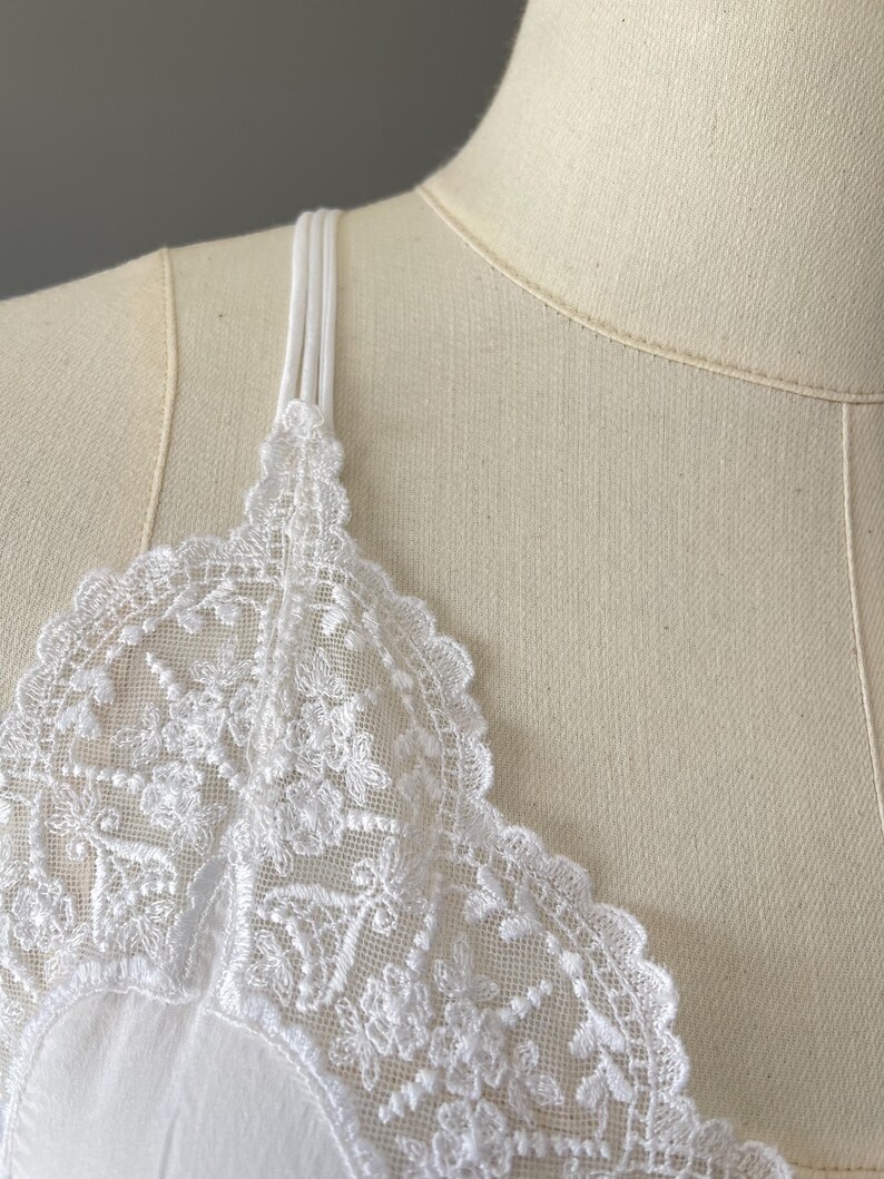 May include: A white camisole featuring delicate floral lace trim along the neckline and thin, triple-strand spaghetti straps. The garment is displayed on a beige mannequin, highlighting the intricate embroidery and scalloped edges of the lace.