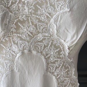 May include: A white garment featuring an intricate floral lace overlay on the upper section. The lace displays a detailed mesh pattern with scalloped edges, contrasting against the smooth, solid white fabric of the main body of the piece.
