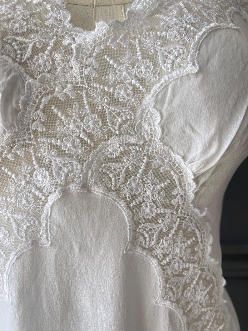 May include: A white garment featuring an intricate floral lace overlay on the upper section. The lace displays a detailed mesh pattern with scalloped edges, contrasting against the smooth, solid white fabric of the main body of the piece.