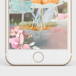 May include: A phone screen displaying a bridal shower invitation with a pastel blue background and a floral design. The text "Lily's Bridal Shower" is displayed on the screen. The image also shows a tiered cake stand with pastries and a floral arrangement.