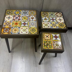 Könnte beinhalten: Drei Holz-Beistelltische mit schwarzen Beinen. Die Tischplatten sind mit bunten, gemusterten Fliesen bedeckt.