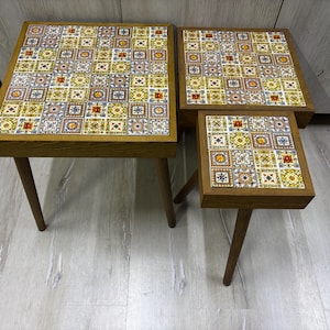 Puede incluir: Juego de tres mesas nido con tableros cuadrados. Las tapas de las mesas presentan un colorido patrón de azulejos en tonos amarillos, azules y naranjas. Las mesas tienen marcos y patas de madera.
