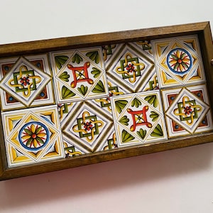 May include: A wooden serving tray with a decorative tile design. The tiles are white with green, yellow, red, and blue geometric patterns. The tray has two brass handles.