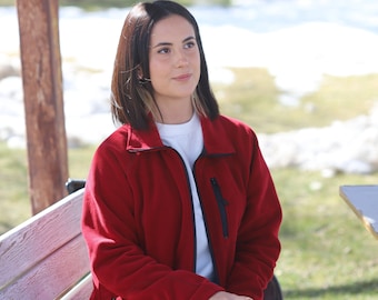 May include: A red fleece jacket with a black zipper and pockets. The jacket is worn over a white t-shirt and paired with black pants. The person is sitting outside on a sunny day.