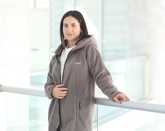 May include: A gray fleece jacket with a hood and a zipper closure. The jacket has a small logo on the chest and a pocket on the left side. The person is wearing a white shirt and dark jeans.