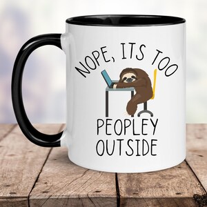May include: White ceramic mug with a black rim and handle. The mug features a cartoon sloth sitting at a desk with a laptop, and the text "NOPE, IT'S TOO PEOPLEY OUTSIDE".