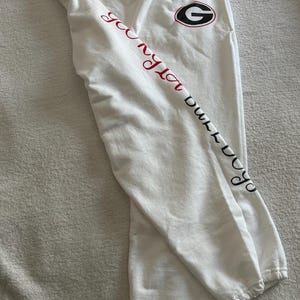 May include: White sweatpants with a drawstring waist and elastic cuffs. The left leg features the text "Georgia Bulldogs" in red and black script, along with a circular logo with a "G" in the center.