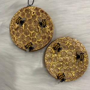 May include: Two round, gold-colored ornaments with a honeycomb pattern and small, black and yellow bees. The ornaments are hanging from a black string.