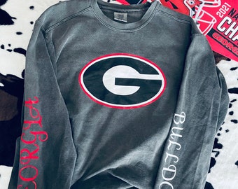 May include: A charcoal gray sweatshirt featuring the University of Georgia Bulldogs logo in the center. The sleeves display "Georgia" in red script and "Bulldogs" in white. A red poster with a football helmet and the words "2021 NATIONAL CHAMPS" is visible.