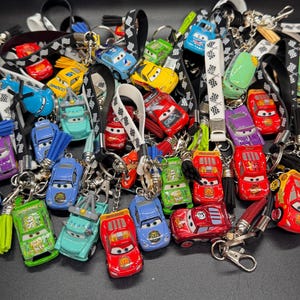 May include: A collection of colorful car-shaped keychains with lanyards. The keychains feature various car designs in red, blue, green, yellow, and purple. The lanyards are black and white checkered, solid white, and black with a silver clasp and tassel.