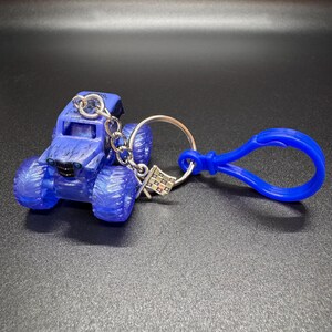May include: A blue monster truck keychain with a silver keyring and a blue clasp. The truck has large tires and a detailed design. A small checkered flag charm hangs from the keyring.