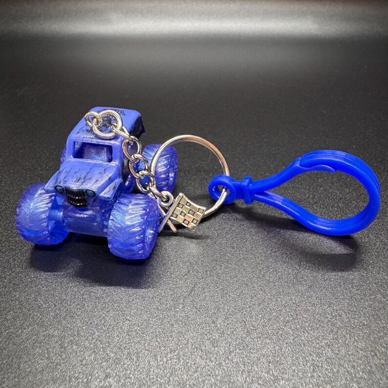 May include: A blue monster truck keychain with a silver keyring and a blue clasp. The truck has large tires and a detailed design. A small checkered flag charm hangs from the keyring.