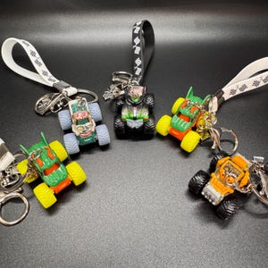 May include: A collection of toy monster truck keychains in various colors, including green, orange, and black. Each keychain features a miniature monster truck with large wheels and a lanyard for easy attachment. The keychains are on a black surface.