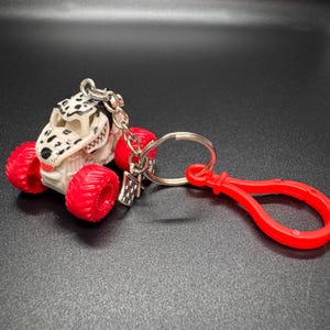 May include: A monster truck keychain with a Dalmatian-themed design. The truck has a white body with black spots, red wheels, and a silver keychain. A red clip is attached to the keychain.