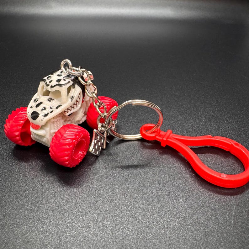 May include: A monster truck keychain with a Dalmatian-themed design. The truck has a white body with black spots, red wheels, and a silver keychain. A red clip is attached to the keychain.