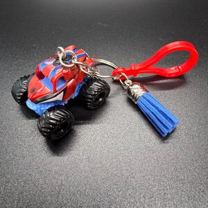 Puede incluir: Un llavero de cami&oacute;n monstruo rojo, azul y negro con un clip rojo y una borla azul. El cami&oacute;n de juguete tiene neum&aacute;ticos negros grandes y un dise&ntilde;o detallado. El llavero est&aacute; sujeto con un anillo plateado.