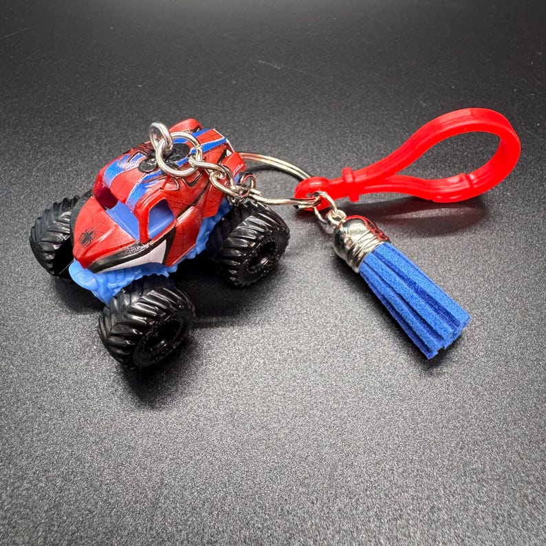 Puede incluir: Un llavero de cami&oacute;n monstruo rojo, azul y negro con un clip rojo y una borla azul. El cami&oacute;n de juguete tiene neum&aacute;ticos negros grandes y un dise&ntilde;o detallado. El llavero est&aacute; sujeto con un anillo plateado.