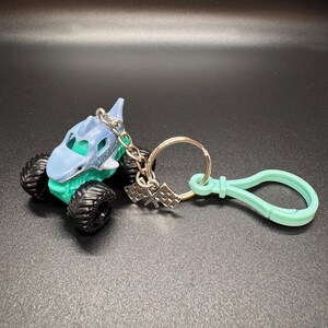 May include: A shark-themed monster truck keychain in light blue and teal. The toy truck has large black tires and a silver chain with a checkered flag charm and a mint green clip.