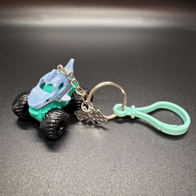May include: A shark-themed monster truck keychain in light blue and teal. The toy truck has large black tires and a silver chain with a checkered flag charm and a mint green clip.