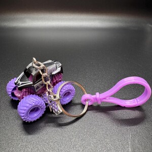 May include: A miniature monster truck keychain with purple wheels and a black and silver body. The keychain includes a silver keyring and a purple clasp.