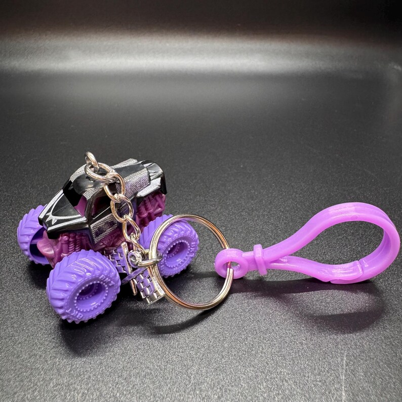 May include: A miniature monster truck keychain with purple wheels and a black and silver body. The keychain includes a silver keyring and a purple clasp.