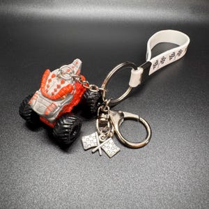 May include: A monster truck keychain with a red and gray body, black tires, and a silver keyring. The keychain includes a white strap with a black pattern and a charm with two checkered flags.