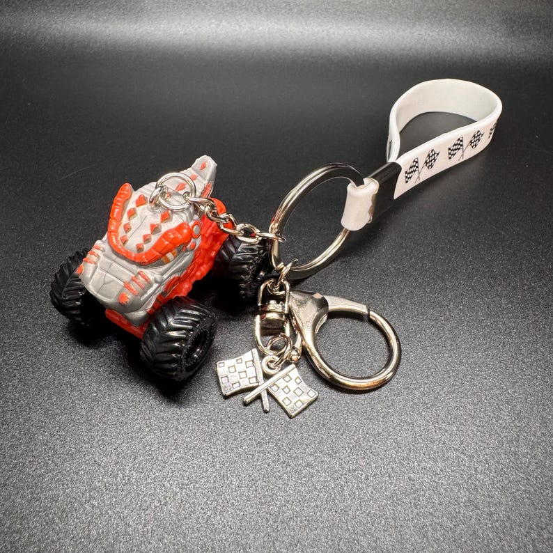 May include: A monster truck keychain with a red and gray body, black tires, and a silver keyring. The keychain includes a white strap with a black pattern and a charm with two checkered flags.