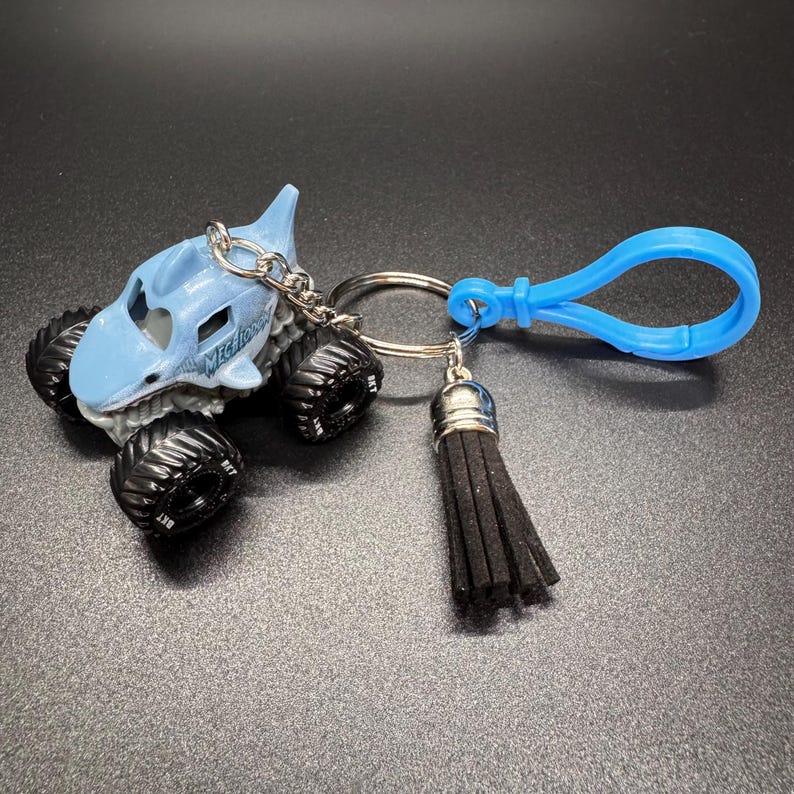 Puede incluir: Un llavero de cami&oacute;n monstruo azul claro con el texto "Megashark" en el lateral. El llavero tiene una cadena plateada, una borla negra y un cierre azul. Los neum&aacute;ticos son negros.