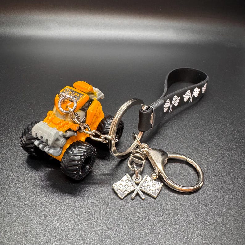 May include: An orange and gray monster truck keychain with a black strap featuring white checkered flags. The keychain includes a silver ring and two silver crossed checkered flag charms. The truck has large black tires.