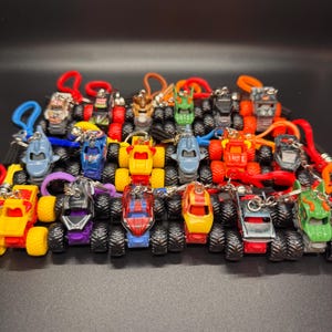 May include: A collection of miniature monster truck keychains in various colors, including red, yellow, blue, and green. Each truck has large, black tires and a unique design. The keychains are attached to colorful loops.