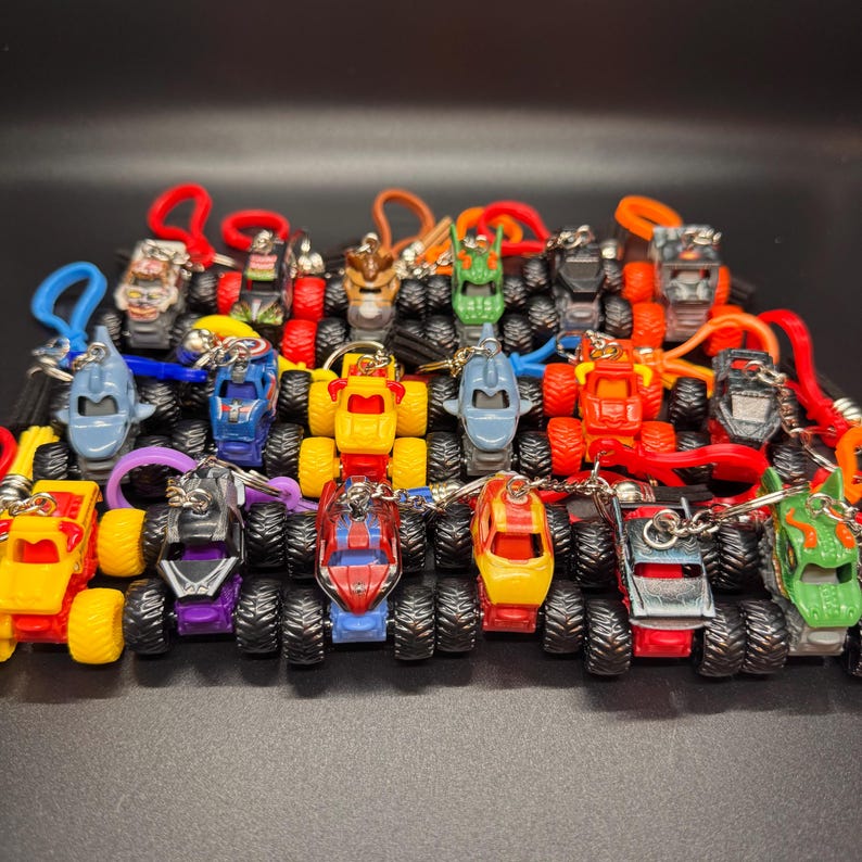 May include: A collection of miniature monster truck keychains in various colors, including red, yellow, blue, and green. Each truck has large, black tires and a unique design. The keychains are attached to colorful loops.
