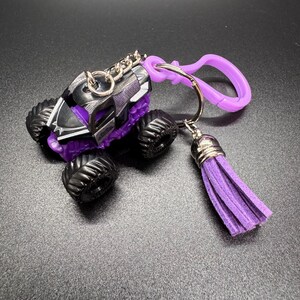 Puede incluir: Un llavero de cami&oacute;n monstruo en miniatura en negro, morado y plateado. El cami&oacute;n tiene neum&aacute;ticos negros grandes y una carrocer&iacute;a detallada. Una borla y un clip morados est&aacute;n unidos al llavero.
