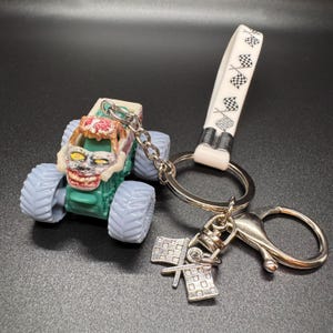 May include: A monster truck keychain with a green body, light blue tires, and a zombie-like face. The keychain includes a silver chain, a white strap with checkered flags, and a silver charm with two checkered flags.