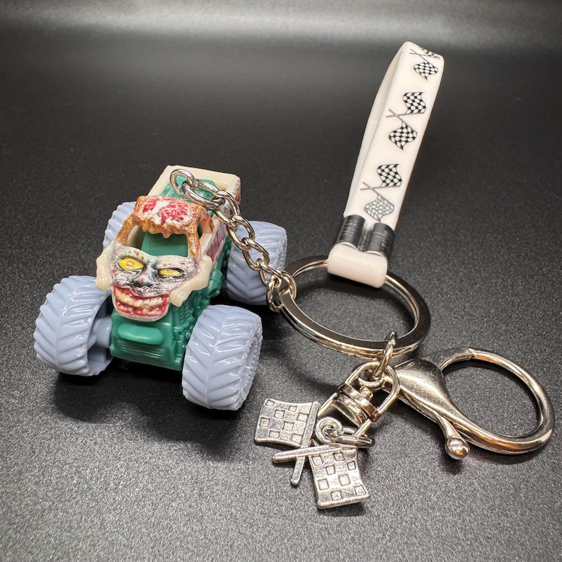 May include: A monster truck keychain with a green body, light blue tires, and a zombie-like face. The keychain includes a silver chain, a white strap with checkered flags, and a silver charm with two checkered flags.