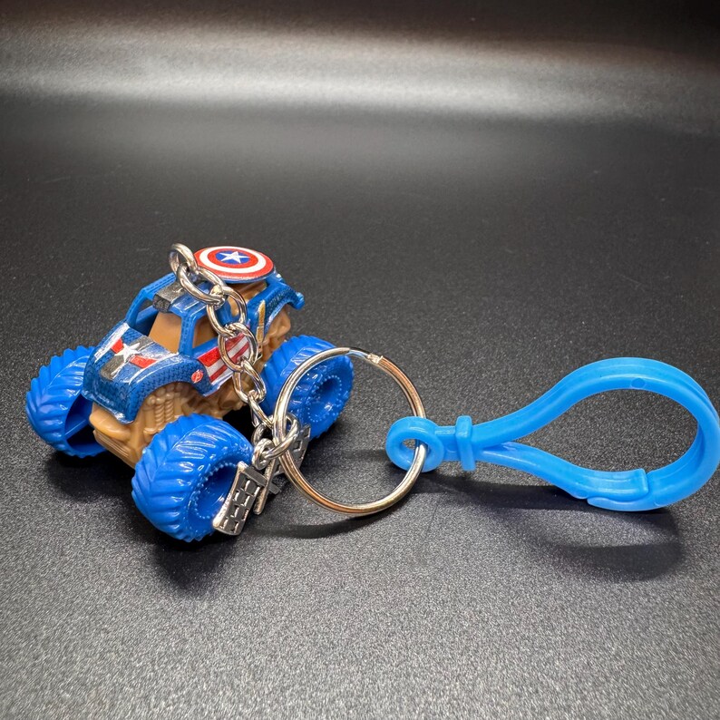 May include: A blue and brown monster truck keychain with a Captain America shield design on top. The truck has large blue tires and is attached to a silver keyring and a blue plastic clip.
