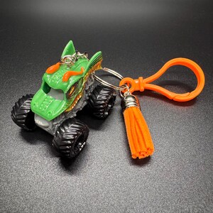 Puede incluir: Un llavero de cami&oacute;n monstruo verde y naranja con un llavero plateado y una borla naranja. El cami&oacute;n tiene ruedas negras y un dise&ntilde;o de drag&oacute;n en la cabina. Un clip naranja est&aacute; unido al llavero.