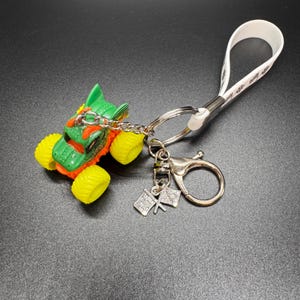 May include: A monster truck keychain with a green and orange body, yellow wheels, and a silver chain. The keychain includes a white strap and a charm with two checkered flags. The background is black.