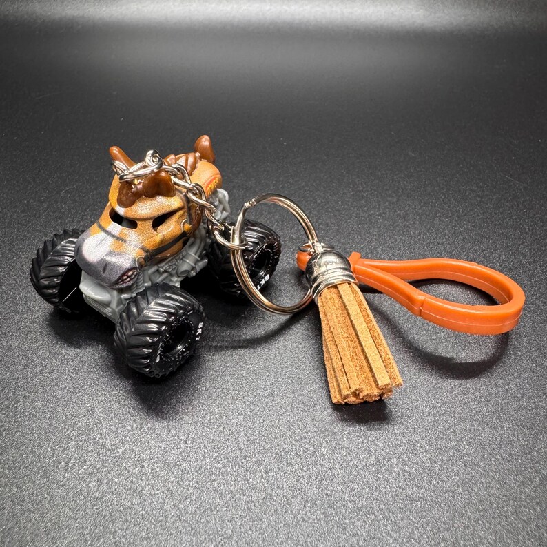 Puede incluir: Un llavero de cami&oacute;n monstruo con dise&ntilde;o de tigre. El cami&oacute;n es naranja, marr&oacute;n y gris con ruedas negras. Tiene una cadena y un llavero plateados, y una borla y un lazo naranjas.