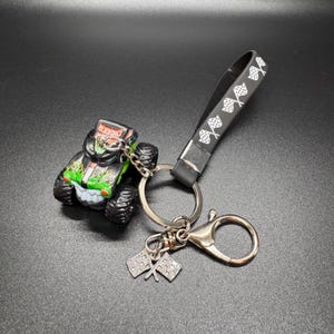 May include: A monster truck keychain featuring a black, green, and red Grave Digger truck. The keychain includes a black strap with white checkered flags, a silver keyring, and two small checkered flag charms.