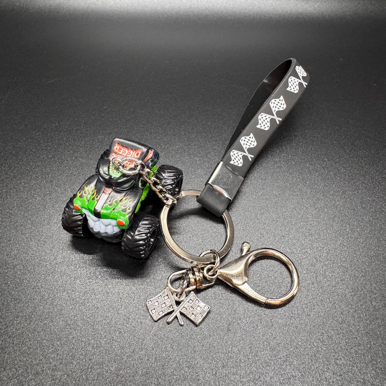 May include: A monster truck keychain featuring a black, green, and red Grave Digger truck. The keychain includes a black strap with white checkered flags, a silver keyring, and two small checkered flag charms.